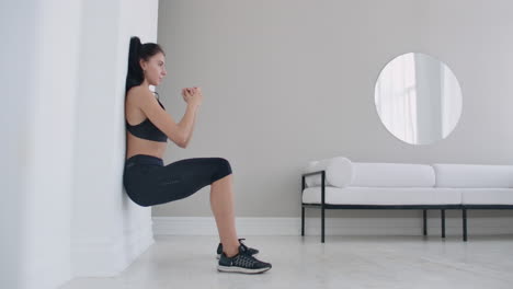 The-brunette-girl-froze-in-a-squat-leaning-on-the-wall-doing-a-static-exercise-in-a-bright-interior.-Keep-90-degree-leg-angle